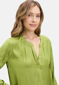 Blusa di seta verde con scollo a V, maniche lunghe fluide e pieghe sottili sulle spalle. Texture liscia con una leggera lucentezza.
