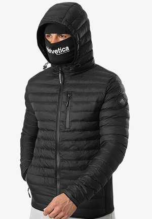 Une personne portant une doudoune noire avec capuche, un bonnet noir et un masque facial noir lisant "Helvetica conçu dans les Alpes."