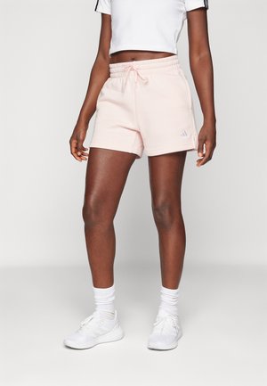 Kvinde iført lyserøde shorts med snor, hvid kort skjorte, hvide sokker og hvide sneakers, stående mod en ensfarvet baggrund.