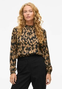 Blusa a stampa leopardata in marrone e nero, tessuto setoso, colletto alto con dettaglio arricciato, maniche lunghe, abbinata a pantaloni neri.