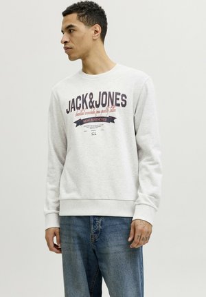 Man draagt een lichtgrijze Jack & Jones trui en blauwe spijkerbroek, staat tegen een effen lichte achtergrond en kijkt opzij.