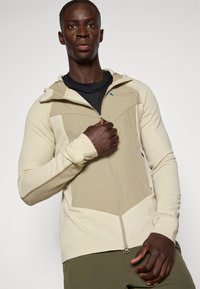 Sweatshirt bege com fecho éclair, fabricado em tecido texturizado, de design ajustado e capuz. Apresenta painéis em contraste e bolsos com fecho éclair.