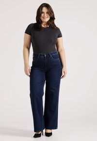 Donkerblauwe high-waisted jeans met een rechte pijp, gemaakt van denim; gecombineerd met een strakke grijze jumpsuit en zwarte hakken.