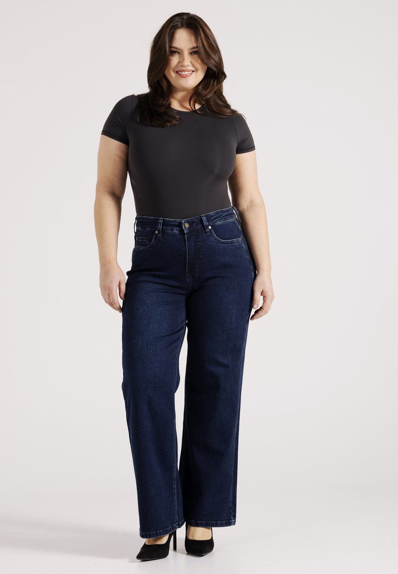 Donkerblauwe high-waisted jeans met een rechte pijp, gemaakt van denim; gecombineerd met een strakke grijze jumpsuit en zwarte hakken.