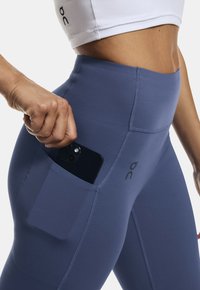 Legginsy z wysokim stanem, niebieskie, wykonane z elastycznego materiału, z boczną kieszenią na smartfona oraz kontrastowymi szwami.