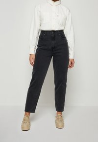 Jeans negros de tiro alto con diseño clásico de cinco bolsillos, combinados con una camisa blanca de botonadura con un bolsillo en el pecho y cuello.