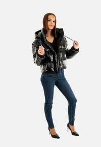 Giacca a vento nera lucida con cappuccio, chiusura con zip e cordini regolabili, abbinata a jeans skinny scuri e tacchi neri.
