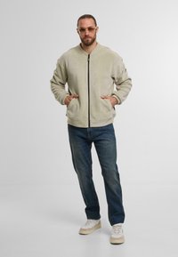 Lichtgroene fleece bomberjack met ritssluiting, zijzakken, gecombineerd met blauwe jeans en beige sneakers, in een minimalistische setting.