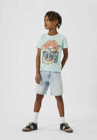 Camiseta verde menta clara con un gráfico de esqueleto, combinada con shorts de mezclilla claros y zapatillas negras y marrones con calcetines blancos.