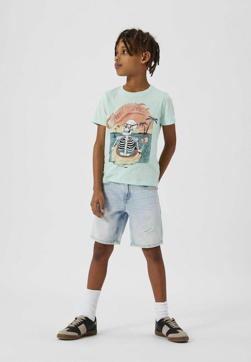 Camiseta verde menta clara con un gráfico de esqueleto, combinada con shorts de mezclilla claros y zapatillas negras y marrones con calcetines blancos.