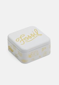 Scatola quadrata bianca in latta con branding giallo e illustrazioni, che presenta la scritta "Fossil" in corsivo e vari design a tema viaggio sui lati.