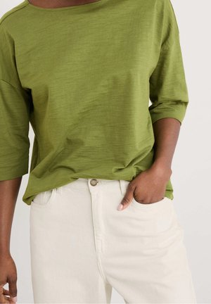 Personne portant une chemise verte à manches trois quarts et un pantalon blanc, une main dans la poche, sur un fond uni.