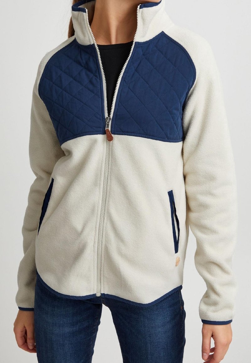 Femme portant une veste polaire crème avec des empiècements matelassés bleu marine aux épaules, des bordures bleu marine et des poches avant zippées, associée à un jean bleu foncé.