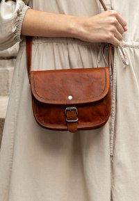 Borsa a tracolla in pelle marrone con pattina, hardware in metallo, tracolla regolabile e dettaglio con fibbia. Texture liscia con accenti di cucitura.