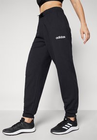 Svarta atletiska joggers med resår i midjan och muddar, med en vit "adidas"-logotyp på vänster ben. Bärs med svarta sneakers.