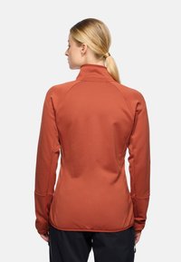 Giacca attiva a maniche lunghe, di colore ruggine, con colletto alto, tessuto testurizzato e maniche raglan. Presenta fori per il pollice e un design aderente.