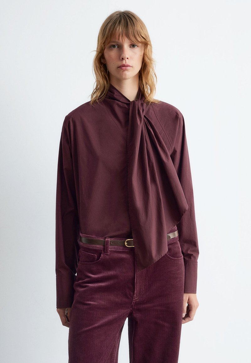 Burgundy shirt with a bow tie detail, long sleeves, and a loose fit, paired with matching corduroy pants and a brown belt.