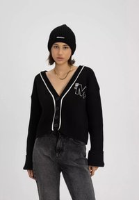 Czarny sweter z dzianiny z głębokim dekoltem w serek, białym wykończeniem, sześcioma guzikami i haftowanym logo. W połączeniu z czarnymi dżinsami i dzianinową czapką.