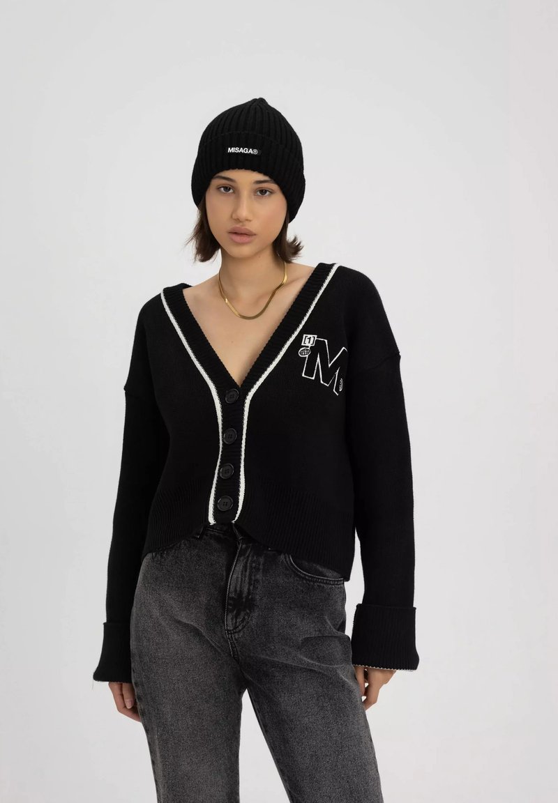 Czarny sweter z dzianiny z głębokim dekoltem w serek, białym wykończeniem, sześcioma guzikami i haftowanym logo. W połączeniu z czarnymi dżinsami i dzianinową czapką.