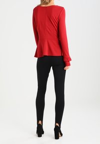 Punainen peplum-paita pitkillä hihoilla, teksturoitu kangas ja takasauma, yhdistettynä mustiin tiukkoihin legginsseihin ja mustiin korkeakorkoisiin kenkiin.