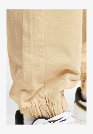 Pantalonsbeige clair avec un motif de grille texturé, dotés d'une taille élastique. Associés à des chaussures de sport noir et blanc.