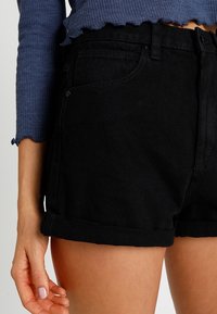 Femme portant un short en denim noir avec ourlets retroussés et un pull court côtelé bleu, main détendue le long du corps.