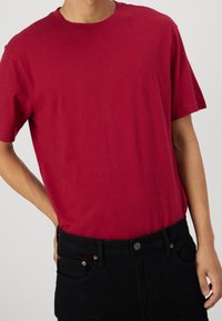 Camiseta de algodón roja con cuello redondo y mangas cortas, con un logo sutil en el pecho, combinada con pantalones vaqueros de mezclilla negra.