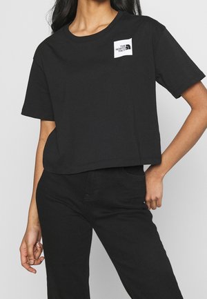 Vrouw in een zwart cropped t-shirt met het The North Face-logo op de borst en zwarte broek, poseert met één hand op de heup.