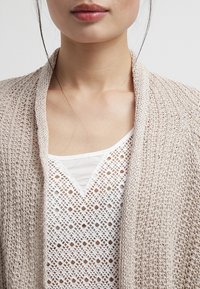 Leichte, beige Strickjacke mit strukturiertem Muster und offenem Vorderteil, über einem weißen Hemd mit diamantförmigen Ausschnitten getragen.