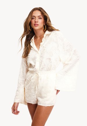 Top blanco texturizado con cuello y mangas largas de campana, combinado con pantalones cortos con cordones a juego. El material es suave y tiene detalles deshilachados.