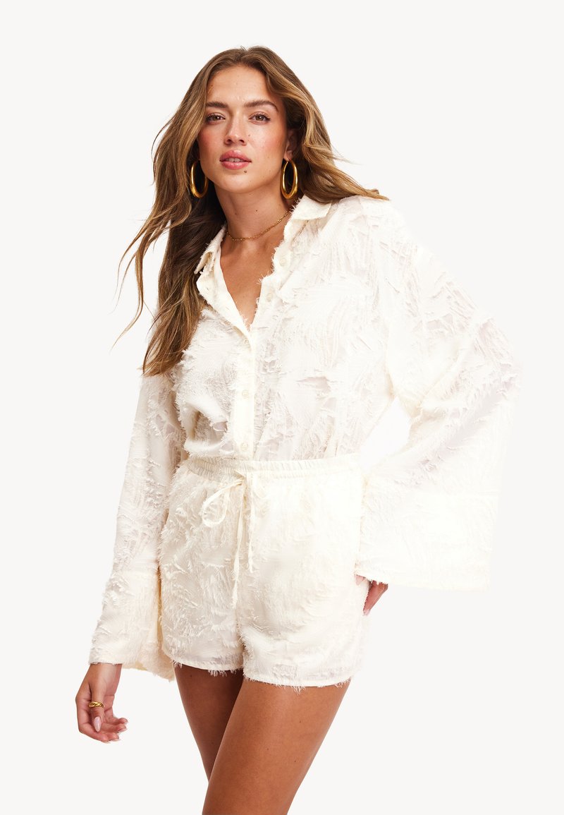 Top blanco texturizado con cuello y mangas largas de campana, combinado con pantalones cortos con cordones a juego. El material es suave y tiene detalles deshilachados.