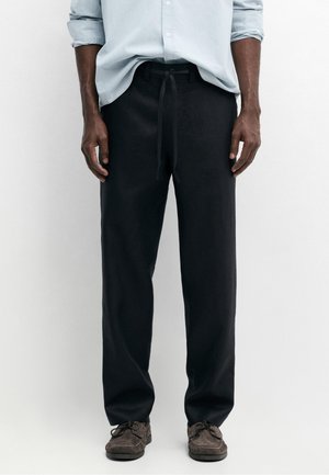Man draagt zwarte broek met trekkoord, lichtblauw overhemd met opgerolde lange mouwen en bruine veterschoenen, staand tegen een effen achtergrond.