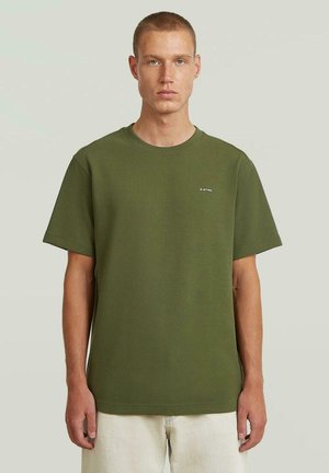 Jeune homme aux cheveux courts portant un t-shirt texturé à manches courtes couleur vert olive et un pantalon de couleur claire, debout devant un fond uni.