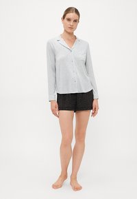 Kvinna som bär en ljusgrå långärmad knäppt pyjamasöverdel och svarta leopardmönstrade shorts, stående barfota mot en enkel bakgrund.