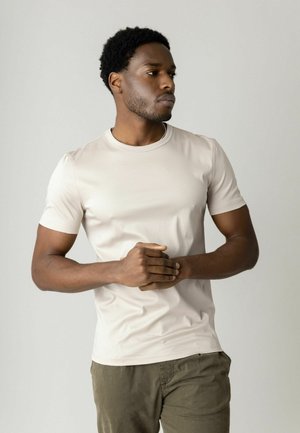 Young man with short curly hair wearing a beige t-shirt and olive pants, standing with hands clasped, looking to the side against a plain background.
