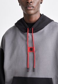 Hoodie gris et noir avec un cordon rouge, présentant un patch logo carré. Le matériau semble doux et texturé avec une coupe ample.