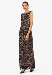 Abito da sera in pizzo nero con motivo floreale su una base nuda, design senza maniche e silhouette lunga fino al pavimento. Indossato con tacchi a spillo neri.
