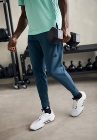 Man i Adidas-byxor och skor som håller en hantel medan han står i ett gym med kettlebells i bakgrunden.