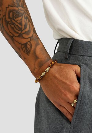 Perlearmbånd med brune og gyldne nuancer, metaliske accenter og et tekstureret design. Båret på et håndled sammen med en ring.