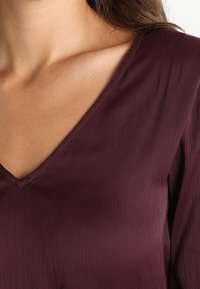 Acercamiento de una persona que lleva una blusa de manga larga de cuello en V de color púrpura oscuro con una sutil textura acanalada y cabello castaño suelto.