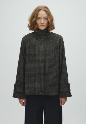Femme aux cheveux bouclés mi-longs porte un manteau boutonné à col montant à carreaux gris foncé et un pantalon noir, debout devant un fond clair uni.