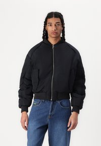 Black puffer jacket with a zip front, ribbed cuffs, and hem. Features two side pockets and a smooth texture. Worn with blue jeans.