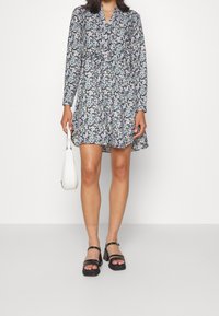 Vestido de manga larga con estampado floral oscuro y cuello; tejido ligero, hasta la rodilla, combinado con sandalias negras de tacón cuadrado y un bolso blanco.