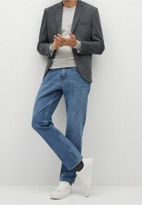 Un uomo indossa un blazer grigio, un felpa leggera grigia, jeans blu e sneakers bianche, in piedi con le gambe incrociate contro un muro bianco.