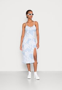 Vestido azul claro com padrões florais, alças finas e racha lateral. Combinado com ténis brancos e óculos de sol angulares.