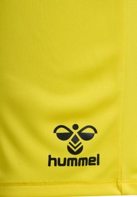 Gul sportströja i mjukt material med en svart "hummel"-logotyp med en symmetrisk design i det nedersta vänstra hörnet.