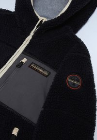 Veste en polaire marine avec une surface texturée et douce. Elle est dotée d'un panneau gris, de deux poches zippées et de patchs de logo de couleurs contrastées.
