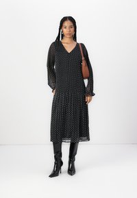 Svart klänning med polkadots, långa transparenta ärmar och V-ringad hals, flödande kjol, parat med knähöga svarta stövlar och en brun handväska.
