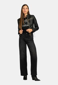 Giubbotto in pelle nera con cerniere, t-shirt grafica nera, jeans larghi scuri e stivaletti neri con tacco. Design semplice e moderno.