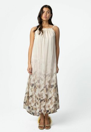 Kobieta ubrana w kremową, bezrękawną sukienkę maxi z falbankowym dekoltem i brązowym kwiatowym wzorem przy brzegu, stojąca na białym tle.